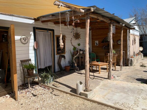 Photo of the exterior of the home and studio of Daniel Rios Rodriguez. Image shows carious styles of tables and chairs, plants, wind chimes, and hanging sculptures