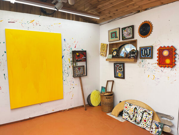 Photo of the studio of Daniel Rios Rodriguez. The photo shows a large yellow painting in progress with various sizes of smaller paintings on the wall adjacent