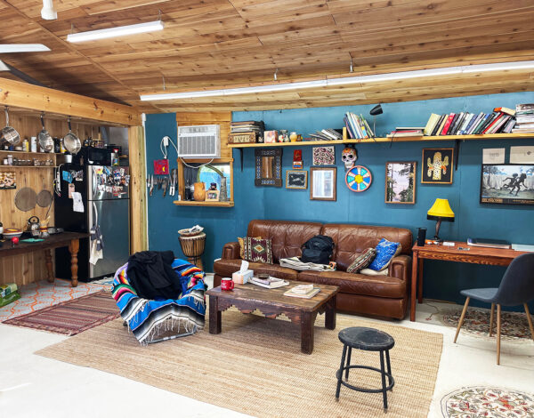 Photo of the studio of Daniel Rios Rodriguez. The image shows a living area with a leather couch and chair covered by a sarape as well as a refrigerator and a high shelf with books.