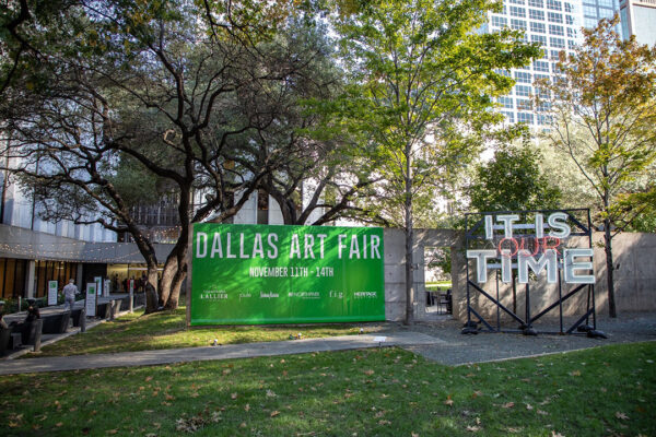 A photograph of the 2021 Dallas Art Fair banner next to a sculpture by Alicia Eggert. The sculpture is a neon work with the words, "IT IS OUR TIME." Photograph by Exploredinary.