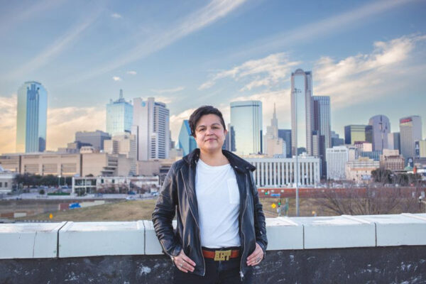 A photograph of artist Angela Faz. They are standing with the Dallas skyline in the background.
