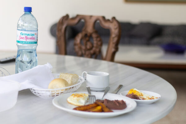 Mesa con desayuno en un patio al aire libre muestra distintos platos con fríjoles, huevos, pan y una botella plástica de agua