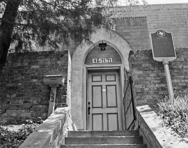 A black and white photograph of the outside of the a building, El Sibil which was the studio of Frank Reaugh. The image shows a staircase that leads to the doorway which is framed by a pointed archway. To the right of the door is a historical marker.