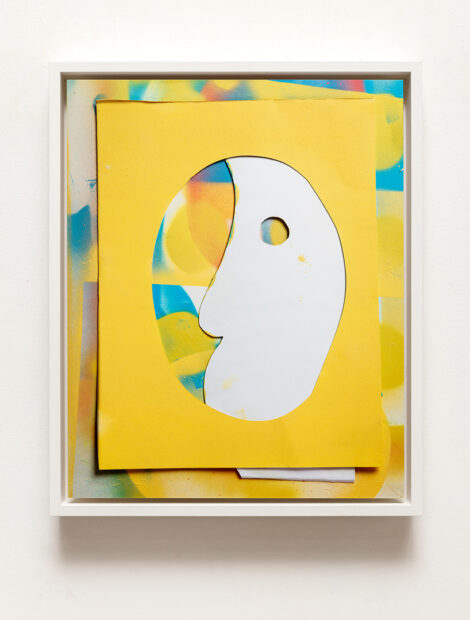 A photograph by Kevin Todora. The photograph is of a piece of white paper cut in the shape of an abstracted profile face. The paper is behind a yellow piece of paper with an oval cut in the center. These two cut paper pieces are arranged in front of a spray painted background that has colors of yellow, blue, red, and green.