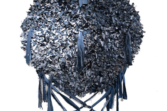 A photograph of a sculptural artwork by Amanda Fay. The work is made from strips of denim that have been arranged to create a large circular shape with longer denim tassels attached.
