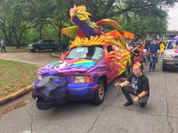 A photograph of a small van that has been transformed into an art car that features a Phoenix -like creature on the roof.