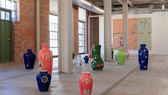 Installation view of large colorful vases with a large painting hanging on a white wall in the background