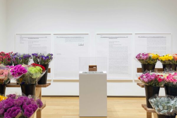 Installation view with flowers on both sides of the frame, a pedestal with box in the enter, and large drawings on the wall behind.