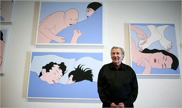 A photograph of artist John Wesley standing in front of three large paintings. He wears all black and holds his hands in front of him as he looks off into the distance. The paintings behind him each include two figures in bed. The figures are painted pink and set against pastel blue sheets.