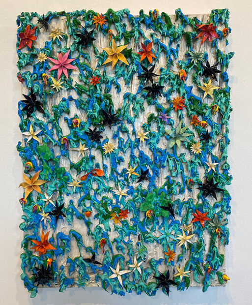A large painting by Julon Pinkston that is filled with thick and messy applications of blue and green paint set against an off-white background. On top of the thick paint blobs are dozens of flowers crafted from dried paint. The flowers vary in size and color.