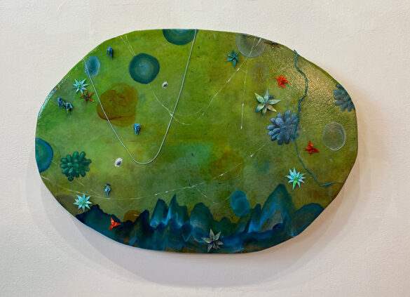 On a large irregular oval shaped canvas, Julon Pinkston has painted an abstract landscape. The bottom of the painting has blue strokes that resemble small mountains or hills. In the upper area are circles and succulent shaped designs painted in blues, greens, and whites set against a greenish brown background. Overlaid on top of the painting are a few strands of blue thread and nearly a dozen flowers sculpted from dried paint.