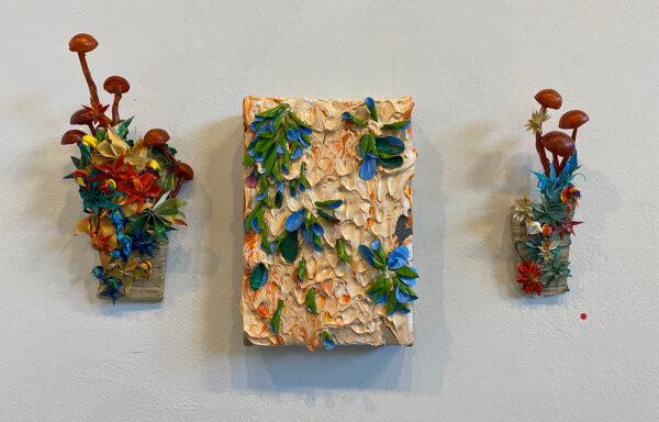 A photograph showing three works by Julon Pinkston hanging on a white gallery wall. The work in the middle is a thickly painted canvas with a light orange background and green and blue petals constructed from dried paint. Flanking the canvas are two wooden vase-like objects with dimensional red mushrooms coming out of them and a slew of various colored flowers constructed from dried paint on top of the vases.