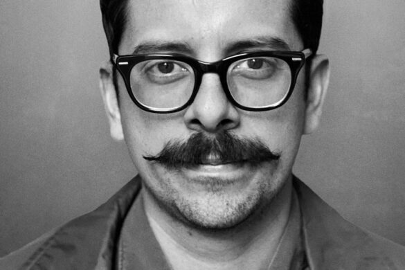 A black and white headshot of Kevin Muñoz. He has short dark hair and a full dark mustache. He wears dark glasses and a button-up shirt. Kevin looks straight into the camera with a slight smile.