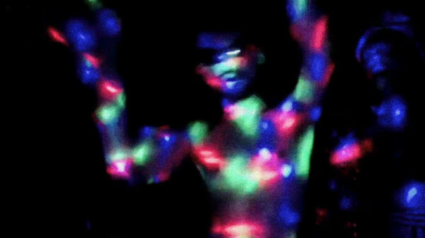 A dark photograph of a a figure dancing with their arms in the air. The figure is illuminated by an array of blue, green, and red lights.