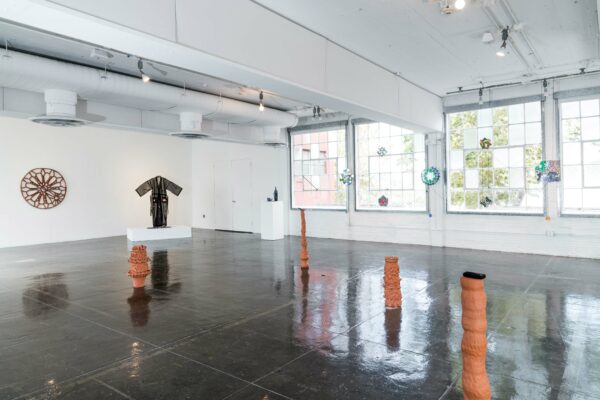 Exhibition view with sculptural work on the floor in the foreground, and a black sheer robe in the background with a rose window made of drain pipes hanging on the wall in the back.
