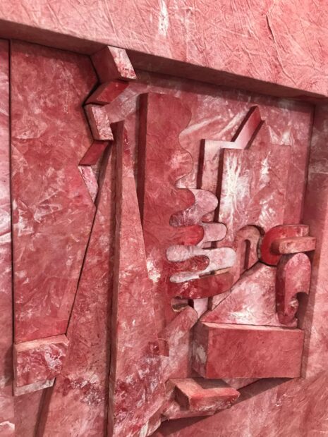 Detail of a pink work that resembles granite with shapes and organic forms protruding in low relief.