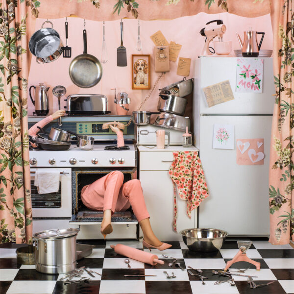 Image of a woman multi-tasking through the oven in a chaotic kitchen. The woman's head and torso are in the oven and her arms reach through the burners holding objects like the phone and kitchen utensils, her legs hang out of the oven.