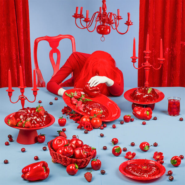 Image of a woman with her head down sitting at a table. She wears all red, including a bright red wig, and is surrounded by red objects and red jello on red platters. The table and background is a contrasting light blue.