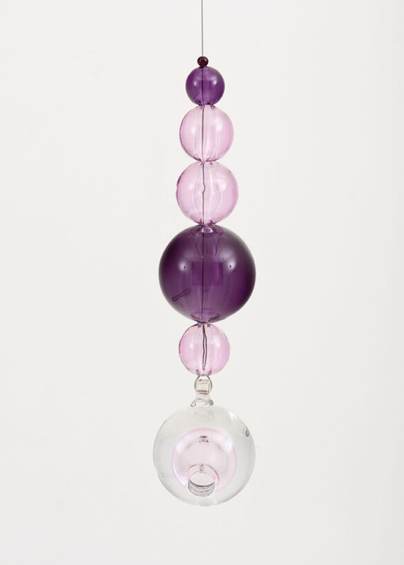 A photograph of a hanging glass sculpture by Jean-Michel Othoniel. The sculpture includes seven spheres of varying sizes. Three of the orbs are a deep purple, three are pink, and one is clear.