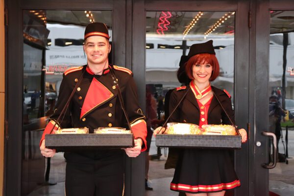 A photograph of a man and a woman each dressed as movie ushers in black, red, and gold outfits. Each person holds a tray of popcorn.