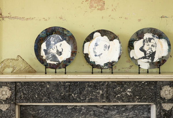 An installation image of three ceramic plates on plate stands sitting on top of a mantel. Each plate has a base of multicolored darkly painted abstract shapes. The center of each plate has been messily painted white and has a depiction of a young man on it. 