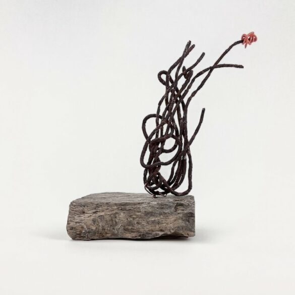 A photograph of a found object sculpture by Sarah Fagan. The abstract sculpture uses thin pieces of metal which are twisted and sit on the right side of the stone base. Atop one piece of brownish metal is a bright red metal.