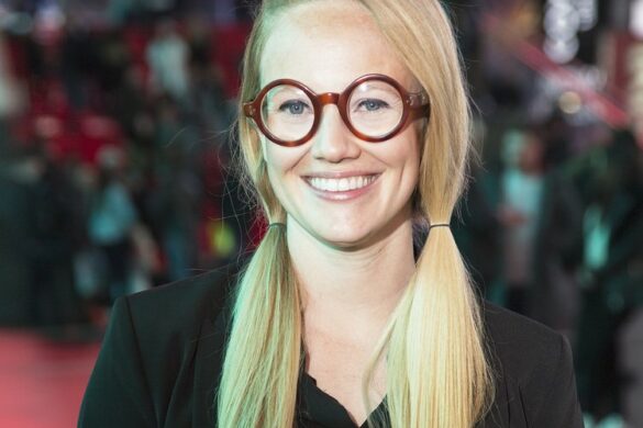 A photograph of VLM (Virginia L. Montgomery). Her long blonde hair is arranged in two ponytails. She wears red circular glasses and a black top. VLM looks into the camera with a full smile.