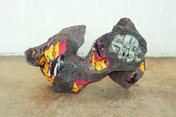 A photograph of a sculpture made from salvaged materials. The sculpture is mostly gray with a rough texture. Some areas of color featuring yellow, red, and black show through and seem to have a smoother texture than the rest of the object. Artwork by Vinchen.