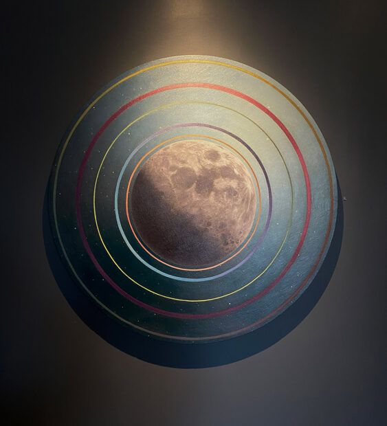 A painting on a circular canvas by Adam Fung. At the center of the canvas is a detailed painting of the moon. It is surrounded by a dark blue sky and dots of distant stars. On top of the sky are five painted rings that seem to emanate from the moon. Each ring is a different color and thickness. 