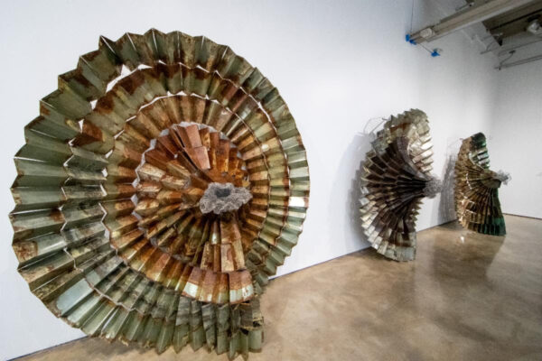 A circular sculpture leaning up against a wall. The piece looks like it is made of metal that has been fanned out.
