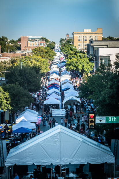 A photograph of the Arts Goggle Festival on Magnolia Street in Fort Worth. The image shows blocks of temporary tents set up and large crowds of people in attendance.