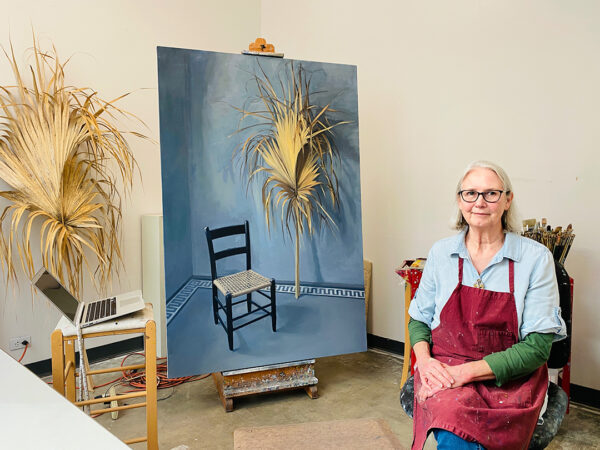 A photograph of artist Carol Ivey sitting in her studio. A large-scale painting sits behind her.