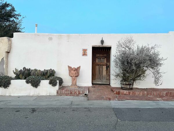 Facade of the Casa Otro Residency in Mesilla, New Mexico. The photo shows a white adobe house with a recessed door, and a terra cotta statue on an angel on the left, and a large succulent plant on the left