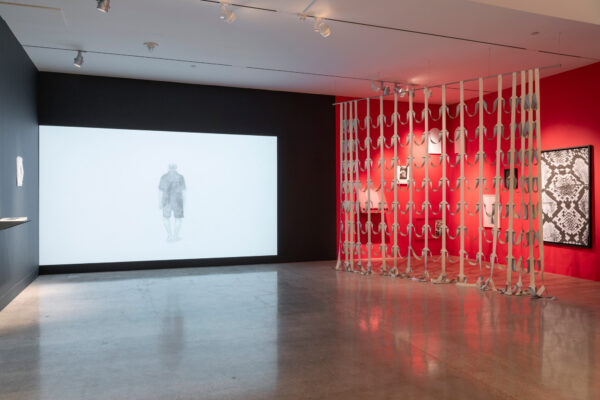 Installation image of a video projected on a black wall, and work hanging on a red wall on the right