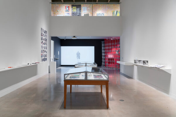 Installation view with glass vitrines in the center of a white walled gallery and shelves on both walls