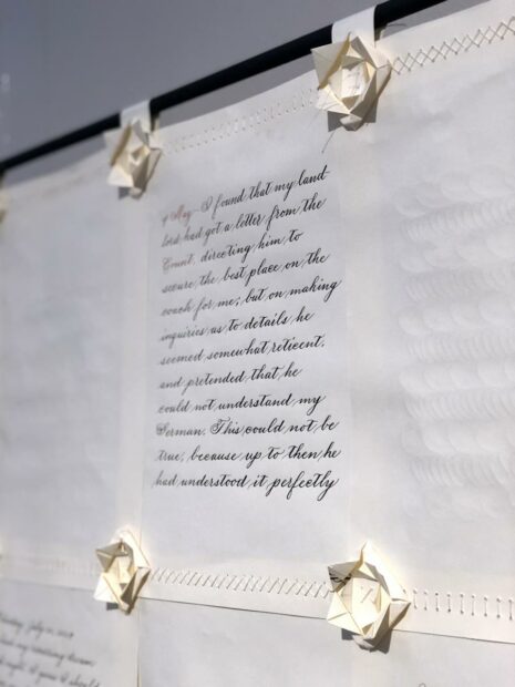 A photograph of a detail of a work of art by Andrea Tosten. The image shows text written in calligraphy on a piece of paper that appears to be sewn together with other pages to make a large document. Origami flowers are affixed to each of the corners.