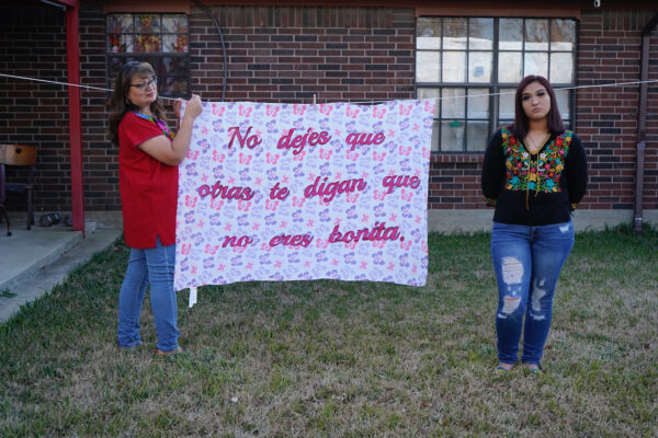 Two women stand on both sides of a sheet hanging on a clothesline. The sheet has the phrase "No dejes que otras te digan que no eres bonita," on it.