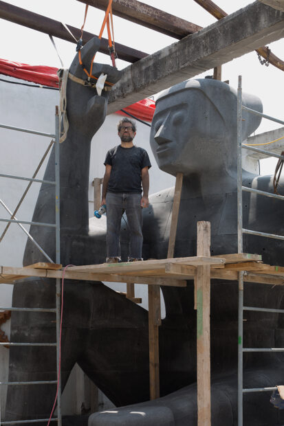 A photograph of artist Pedro Reyes standing on scaffolding next to his large-scale sculpture, titled "Stargazer (Citlali)."