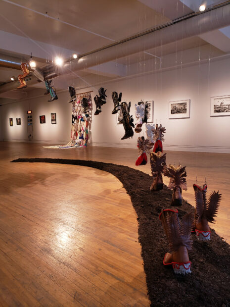 Installation view of boots rising to the ceiling, works on paper on the walls in the gallery