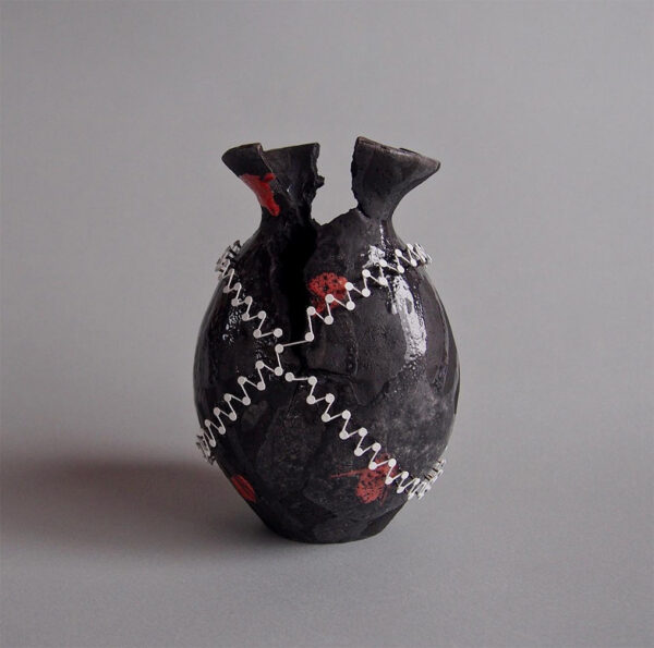 A photograph of a ceramic object made by Keita Suto. The sculpture is of a sake bottle which appears to be broken at the top. The bottle is painted black and has white nails sticking out of it. The nails form an X pattern across the object and are connected by a white thread. There are also spots or red paint on the vessel.