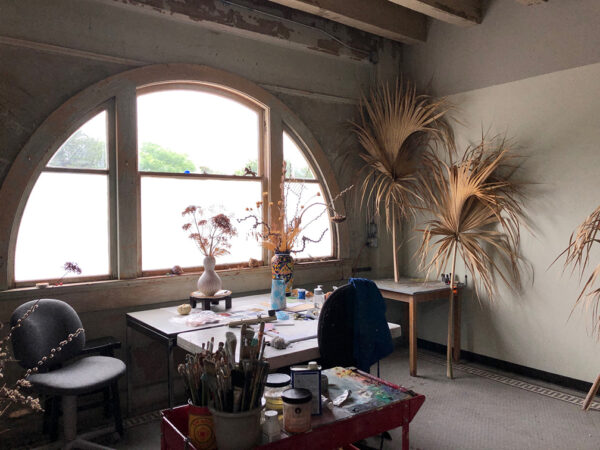 A photograph of the inside of an artist studio. There is a arched window that lets in a substantial amount of light. There are also multiple tables each with still life objects like dried plants and vases.