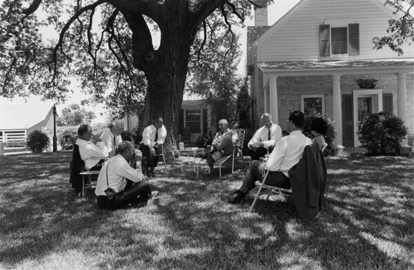 A black and white photograph by Mike Geissinger of President Lyndon B. Johnson holding a meeting with representatives of the press seated in chairs below a large oak tree.