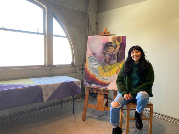 A photograph of Michelle Cortez Gonzales sitting in her studio. A large arched window lets in light to the left and a work of art sits on an easel beside the artist.