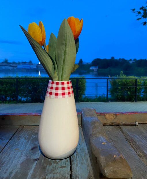 Orange, fabric tulips in a white vase with a band of a red and white checkered pattern on the top. It is a table setting overlooking the Narva River