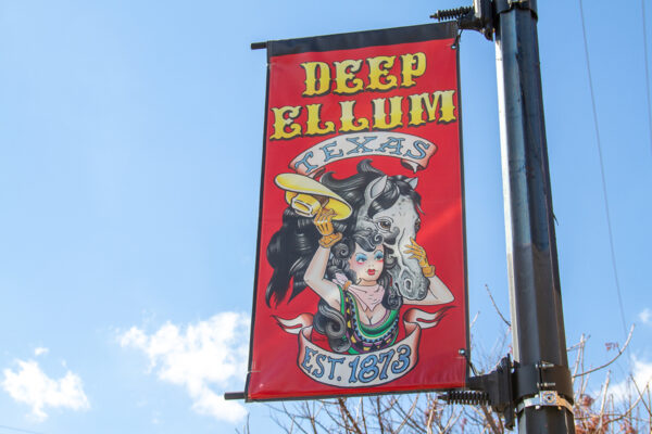 A photograph of a tall narrow banner attached to a street pole. The banner has text that reads, "Deep Ellum Texas, Est. 1873." The image on the banner is of a woman with long curyly black hair and her horse. Design by Norman Dean Williams.