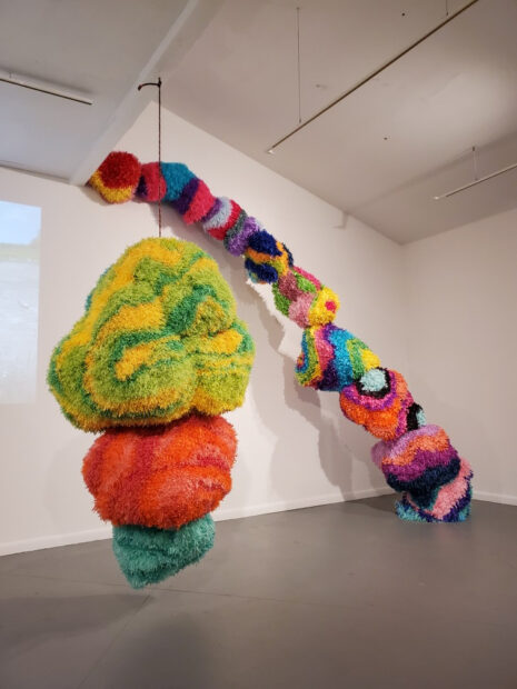 Two large scale sculptures made of piñata paper hanging from the ceiling and leaning agains a wall.