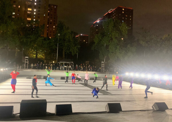 Photo of dancers in multi-colored costumes dancing on an outdoor stage in New York City