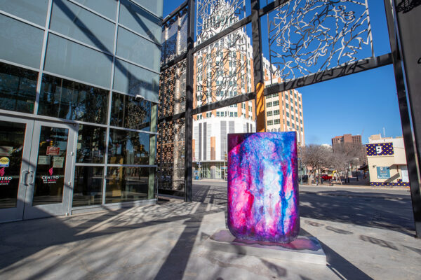 A photograph of a paleta sculpture by David Blancas. The 10-foot sculpture is of an overturned paleta. The paleta is painted with swirls of pink, blue, and purple. 