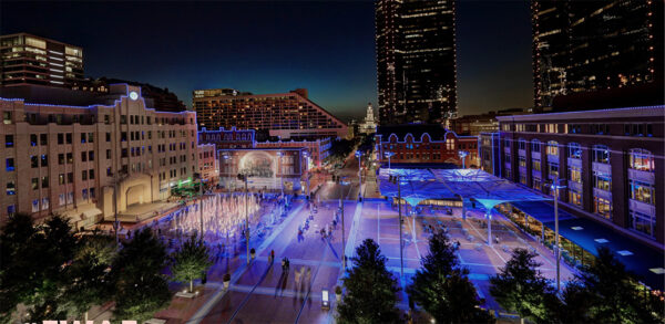 A night time photograph of Sundance Square Plaza in downtown Fort Worth. The image shows a fountain on the right and covered seating on the left with buildings surrounding the plaza.