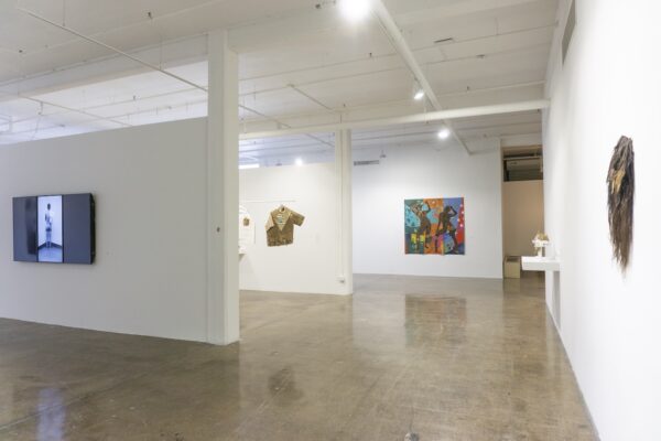 A photograph of a white-walled gallery with a variety of artworks displayed. Artworks include a video on a large flatscreen monitor, clothing items, a large scale painting, and small sculptures on a white shelf.